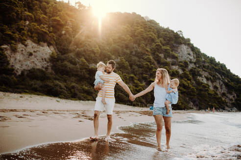 A family with two toddler children walking on beach on summer holiday at sunset. A father and mother carrying son and daughter in the arms, holding hands. - HPIF23795