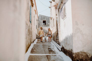 A family with two toddler children walking in town on summer holiday. A father and mother with son and daughter in baby carrier on a narrow street. - HPIF23803