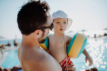 Ein Vater mit einem kleinen Kind mit Armbändern steht am Swimmingpool im Sommerurlaub. - HPIF23809