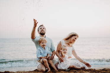 Eine Familie mit zwei Kleinkindern sitzt im Sommerurlaub am Sandstrand und spielt. - HPIF23823