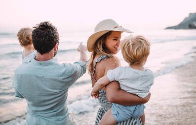 Eine Familie mit zwei Kleinkindern, die im Sommerurlaub bei Sonnenuntergang am Strand spazieren gehen. Ein Vater und eine Mutter tragen Sohn und Tochter auf dem Arm. - HPIF23832
