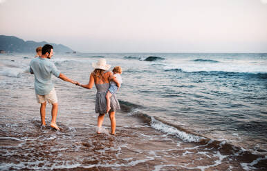 Rückansicht einer jungen Familie mit zwei Kleinkindern, die im Sommerurlaub am Strand spazieren gehen. - HPIF23835