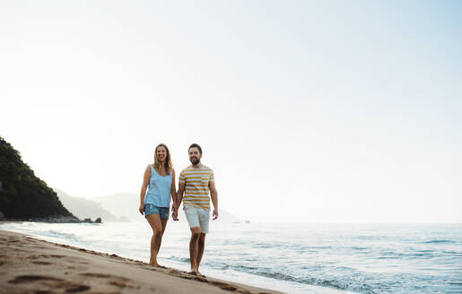 Ein fröhlicher Mann und eine fröhliche Frau, die im Sommerurlaub am Strand spazieren gehen und sich an den Händen halten. Copy space. - HPIF23867