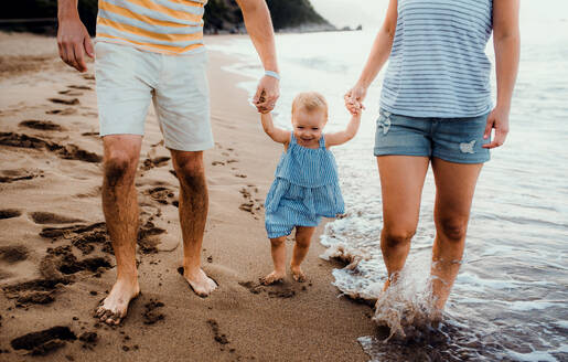 Ein Mittelstück von Eltern mit ihrer kleinen Tochter, die im Sommerurlaub am Strand spazieren gehen und sich an den Händen halten. - HPIF23874