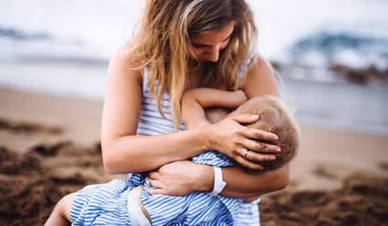 A young mother breasfeeding toddler daughter on beach on summer holiday. - HPIF23881