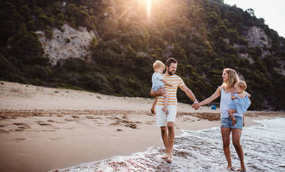 A family with two toddler children walking on beach on summer holiday at sunset. A father and mother carrying son and daughter in the arms, holding hands. - HPIF23892