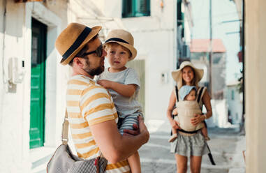 A family with two toddler children walking in town on summer holiday. A father and mother with son and daughter in baby carrier on a narrow street. - HPIF23901