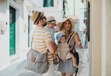 Eine junge Familie mit zwei Kleinkindern und Hüten geht im Sommerurlaub in der Stadt spazieren. - HPIF23902