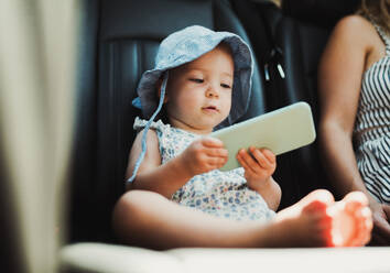 Ein kleines Mädchen im Auto sitzend und mit einem Smartphone spielend. Ein Sommer. - HPIF23914