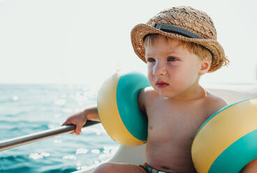 A small boy with armbands sitting on boat on summer holiday, holding a railing. - HPIF23916