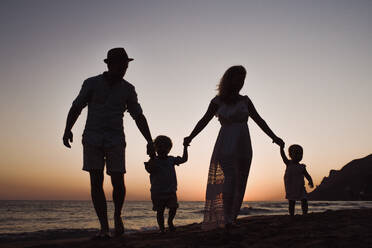 Eine Familie mit zwei Kleinkindern, die in der Abenddämmerung am Strand spazieren gehen, Silhouetten, die sich an den Händen halten. - HPIF23952