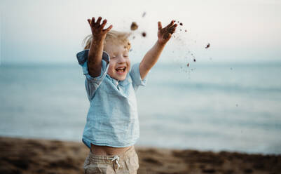A cheerful small toddler boy standing on beach on summer holiday, throwing sand. - HPIF23971
