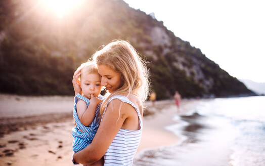 Junge Mutter, die ein kleines Mädchen am Strand im Sommerurlaub bei Sonnenuntergang umarmt. - HPIF23973