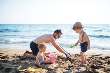 Ein reifer Vater mit zwei Kleinkindern, die im Sommerurlaub am Strand mit Sand spielen. - HPIF23979