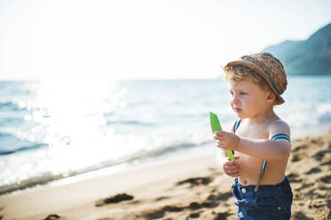 Ein kleiner Junge mit Hut und Shorts, der im Sommerurlaub am Strand steht und einen Spaten in der Hand hält. Copy space. - HPIF23981
