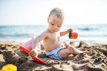 A small topless toddler girl sitting on beach on summer holiday, playing in sand. - HPIF23983