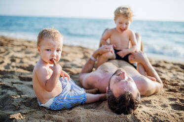 A father with two toddler children lying on sand beach on summer holiday, playing. - HPIF23992