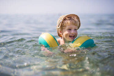 Ein glücklicher kleiner Junge mit Hut und Armbändern, der im Sommerurlaub im Wasser schwimmt, mit Sand auf den Wangen. - HPIF24013