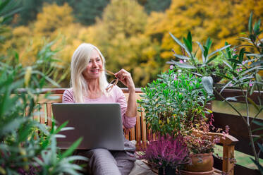 Eine ältere Frau mit Laptop sitzt im Freien auf einer Terrasse und arbeitet. - HPIF24014