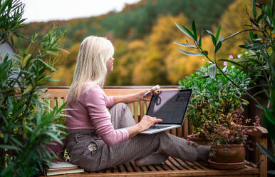A senior woman architect with laptop sitting outdoors on terrace, working. - HPIF24016