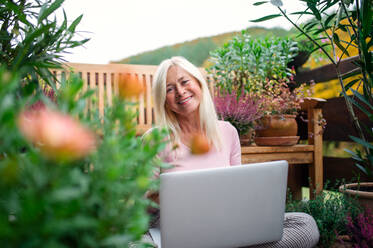 Eine ältere Frau mit Laptop sitzt im Freien auf einer Terrasse und arbeitet. - HPIF24019