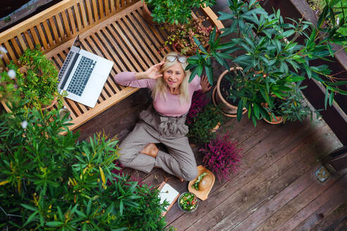 Draufsicht auf eine ältere Frau mit Laptop, die im Freien auf einer Terrasse sitzt und sich ausruht. - HPIF24026