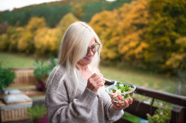 Ältere Frau steht im Freien auf einer Terrasse und isst einen gesunden Salat-Snack. - HPIF24046