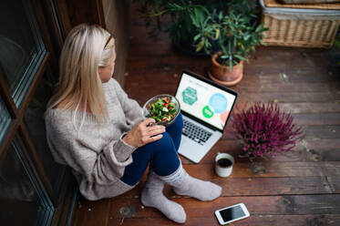 Senior woman with laptop sitting outdoors on terrace, working and eating. - HPIF24047