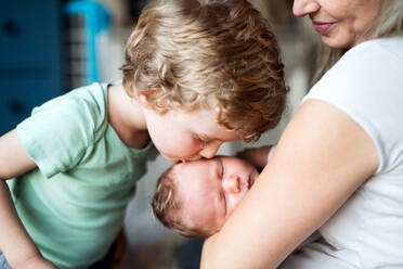 Ein kleiner Junge küsst zu Hause sein schlafendes neugeborenes Brüderchen, die Mutter hält es im Arm. - HPIF24082