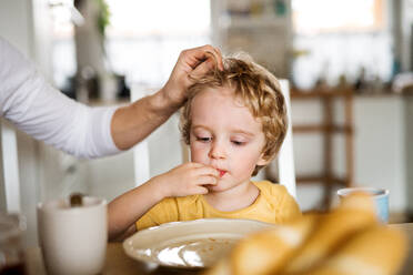 A front view of a sad toddler boy sitting at the table at home, eating. - HPIF24106