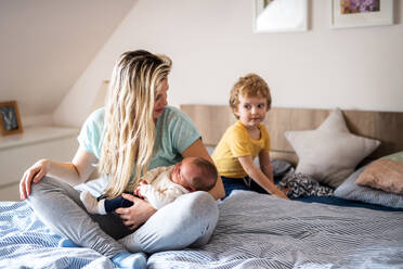 Eine schöne junge Mutter mit einem neugeborenen Baby und seinem kleinen Bruder zu Hause, auf dem Bett sitzend. - HPIF24137