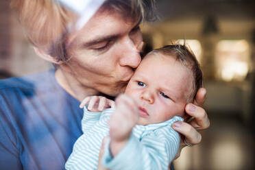 Ein junger Vater hält sein neugeborenes Baby zu Hause im Arm und küsst es, aufgenommen durch Glas. - HPIF24152