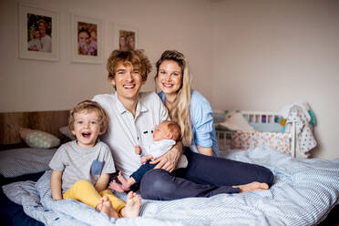 Porträt junger Eltern mit einem neugeborenen Baby und einem kleinen Sohn im Kleinkindalter, die zu Hause auf dem Bett sitzen. - HPIF24197