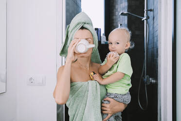In ein Handtuch gewickelte Mutter, die ihren Sohn trägt und vor dem Badezimmer Kaffee trinkt - NDEF00615