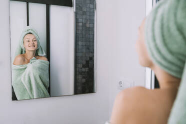 Lächelnde Frau, eingewickelt in ein Handtuch, schaut in den Badezimmerspiegel - NDEF00618