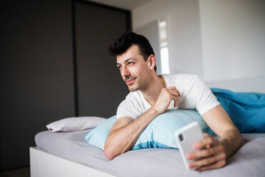 Ein junger Mann mit Smartphone im Bett zu Hause, der eine SMS schreibt. - HPIF24204