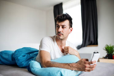 Ein junger Mann mit Smartphone im Bett zu Hause, der eine SMS schreibt. - HPIF24205