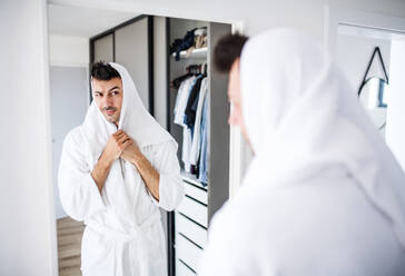 Junger Mann mit Bademantel, der sich im Schlafzimmer die Haare trocknet, eine morgendliche Routine. - HPIF24218