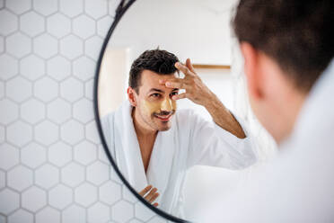 Junger Mann, der sich morgens im Badezimmer eine Maske aufs Gesicht legt, eine tägliche Routine. - HPIF24236