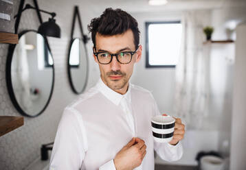 Ein junger Mann mit weißem Hemd steht morgens mit einer Tasse Kaffee in der Hand im Badezimmer. - HPIF24265