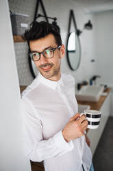 Ein junger Mann mit weißem Hemd steht morgens mit einer Tasse Kaffee in der Hand im Badezimmer. - HPIF24266