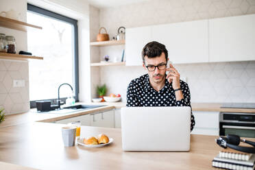 Junger Mann mit Laptop und Smartphone in der Küche sitzend und arbeitend, ein Home Office Konzept. - HPIF24280