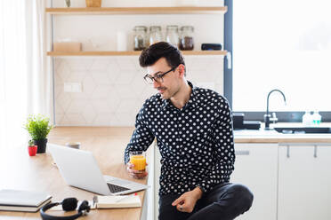 Young man with laptop sitting in kitchen, holding juice. A home office concept. - HPIF24282