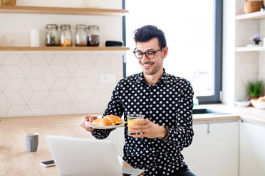 Fröhlicher junger Mann mit Frühstücksset und Laptop zu Hause bei einem Skype-Gespräch. - HPIF24283