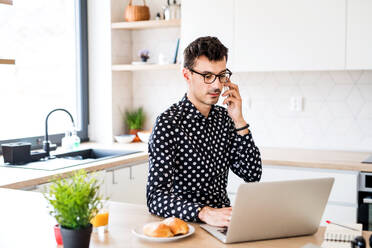 Junger Mann mit Laptop und Smartphone in der Küche sitzend und arbeitend, ein Home Office Konzept. - HPIF24285