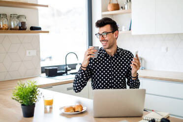 Junger Mann mit Laptop und Smartphone in der Küche sitzend und arbeitend, ein Home Office Konzept. - HPIF24287