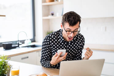 Young man with laptop and smartphone sitting in kitchen, expressing excitement. A home office concept. - HPIF24290