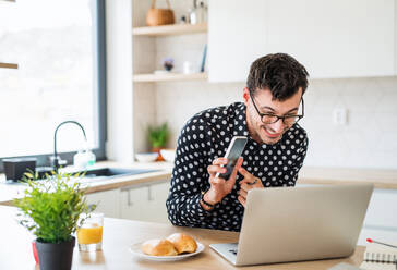 Young man with laptop and smartphone sitting in kitchen, working. A home office concept. - HPIF24291