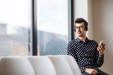 Ein junger Mann mit Kopfhörern und Smartphone sitzt am Fenster und hört Musik. - HPIF24294