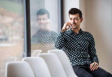 Ein junger Mann mit Smartphone, der am Fenster sitzt und telefoniert. Raum kopieren. - HPIF24298